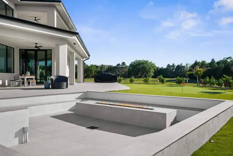 A backyard poolside scene with a clean white patio and a contemporary home in the background under a clear sky.