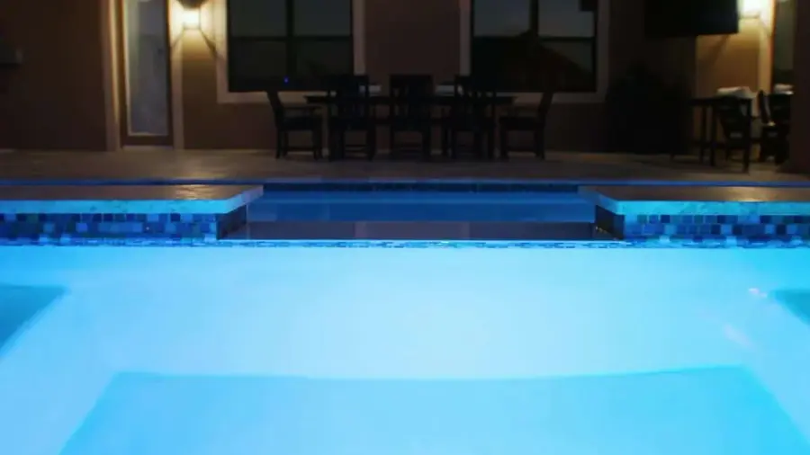A brightly lit swimming pool at night, showcasing a modern pool design with tiled edges and adjacent patio dining furniture.