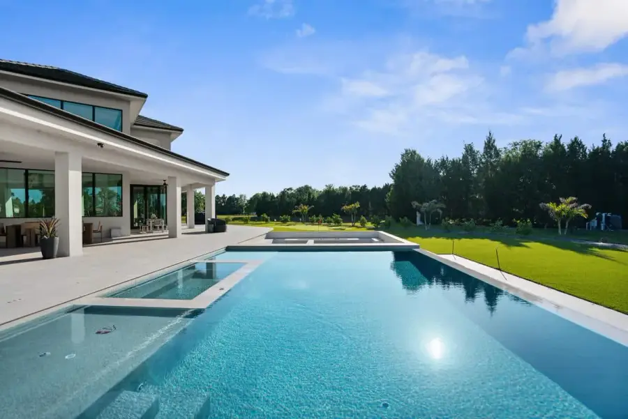 A clear blue swimming pool reflecting the sky, surrounded by a modern house and well-manicured greenery.