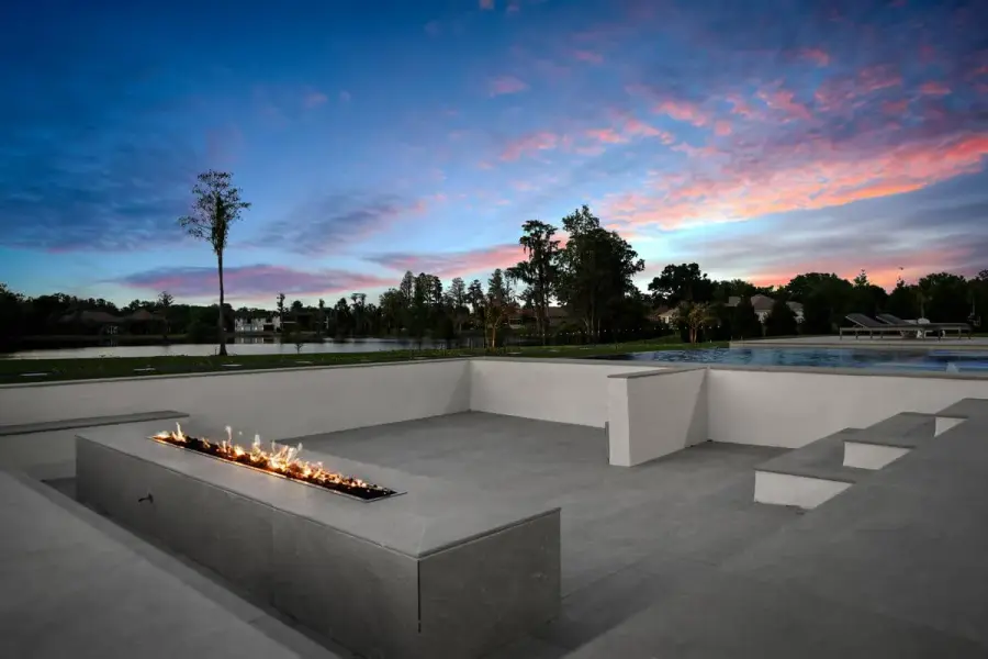 A glowing fire pit surrounded by smooth concrete seating, framed by a modern pool and lush green landscape at sunset.