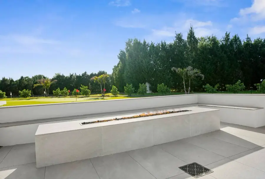 A polished backyard fire feature along a white concrete bench, facing an open lawn with trees in the distance.