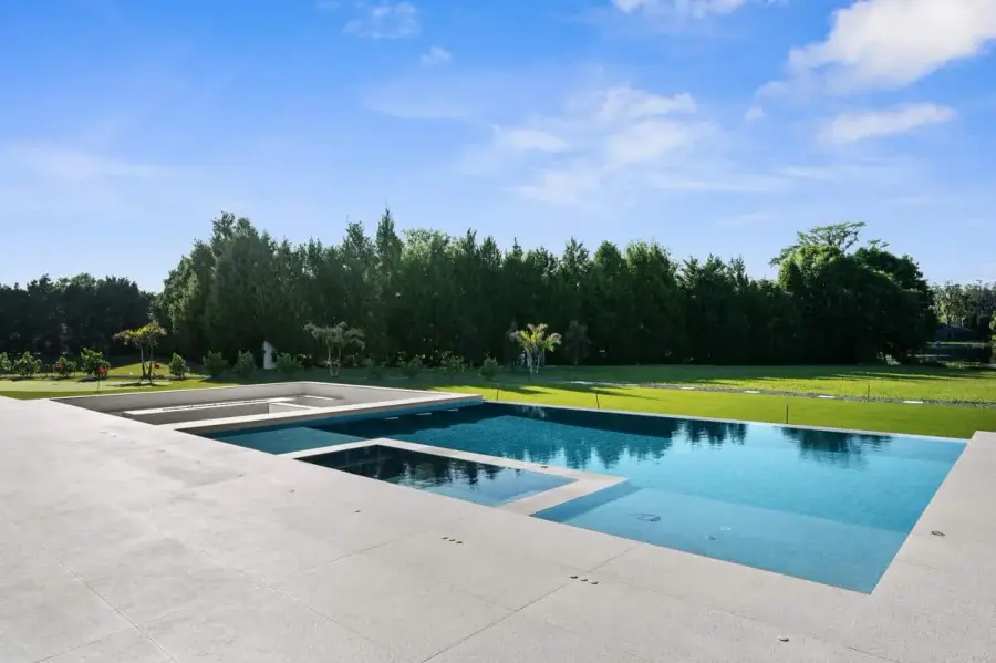 A rectangular pool with crystal-clear water and a modern white patio, surrounded by green trees and a sunny sky.