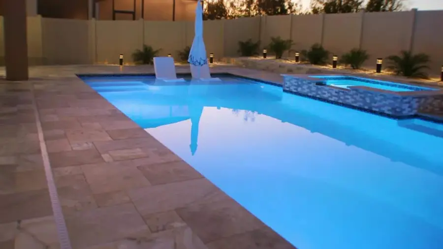 A serene pool with integrated lounge chairs and an umbrella on a pool sun shelf, surrounded by travertine decking.