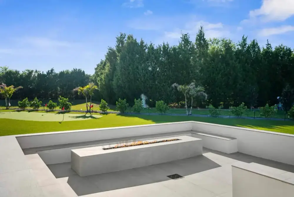 A crisp white concrete bench and fire pit facing a grassy backyard area with tall trees and a peaceful atmosphere.