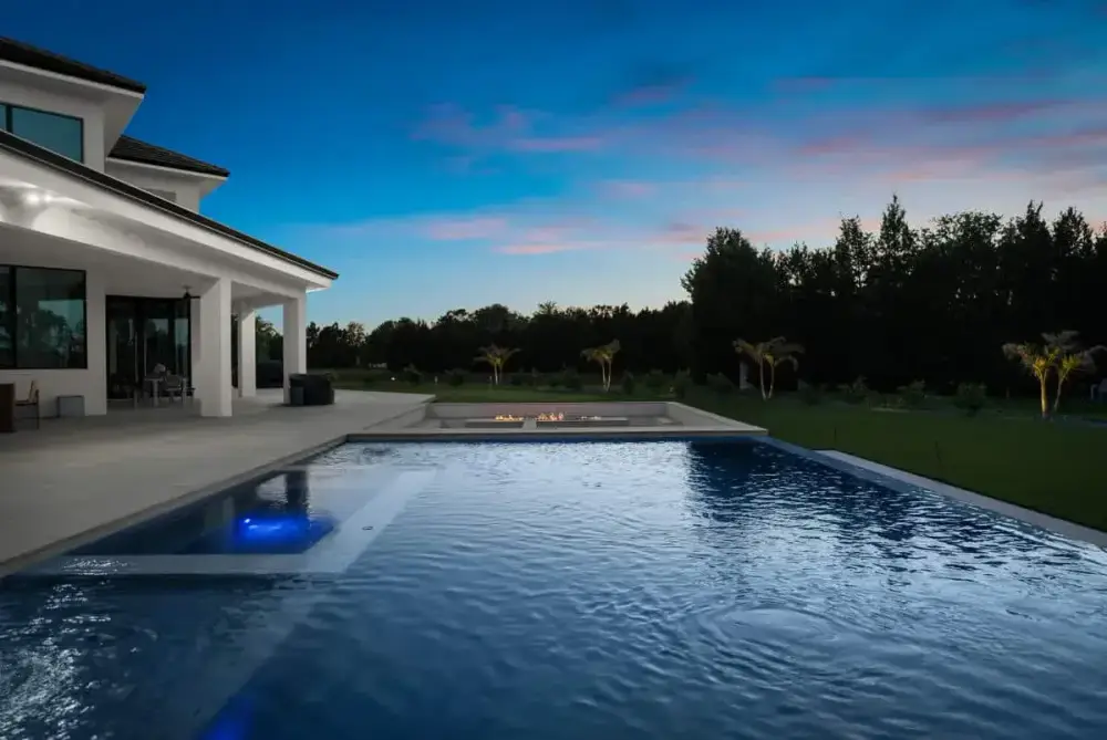 A luxurious modern home with a pool in the foreground, reflecting the warm hues of the sunset sky and surrounded by sleek architecture.