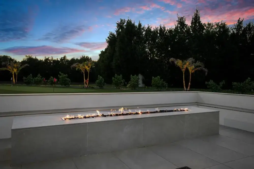 A modern fire feature framed by vibrant green trees and undergrowth, set against the backdrop of a sunset sky.