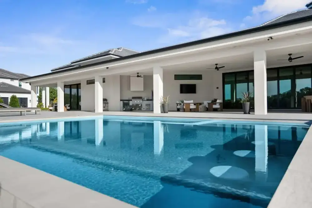 A reflective backyard pool surrounded by white stone decking and a contemporary home with a covered patio.