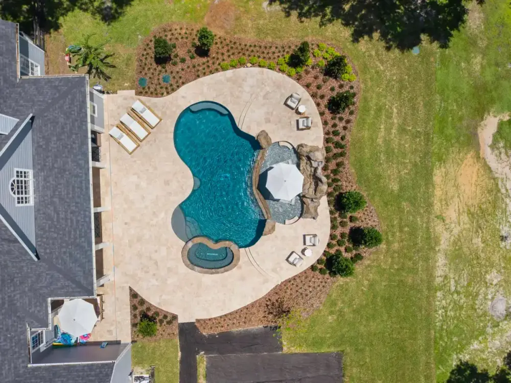 Drone shot of a custom pool with sun shelf, spa, and lounge areas, set in a spacious backyard with landscaped border.