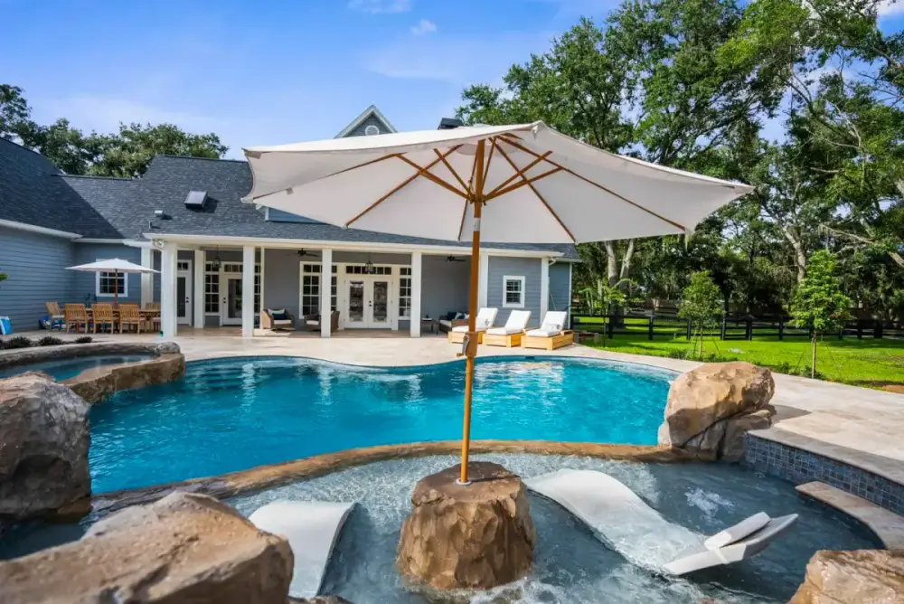 Resort-style pool featuring a built-in Baja shelf with a lounging chair and umbrella, framed by natural rock features and a luxury patio.