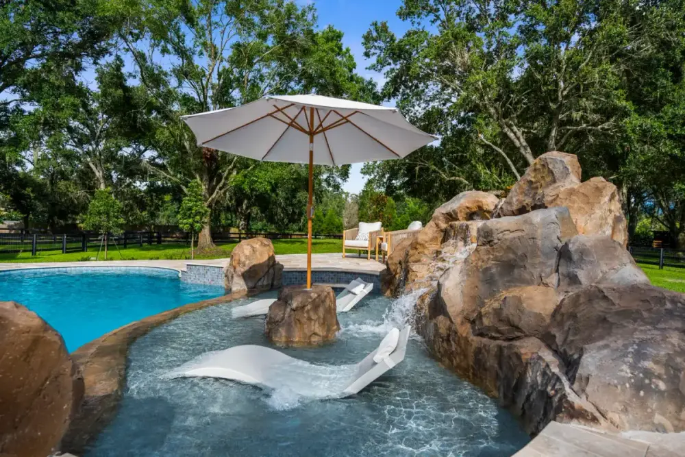 Modern pool design featuring a baja shelf with an in-pool lounger and umbrella, surrounded by natural stone and lush greenery.