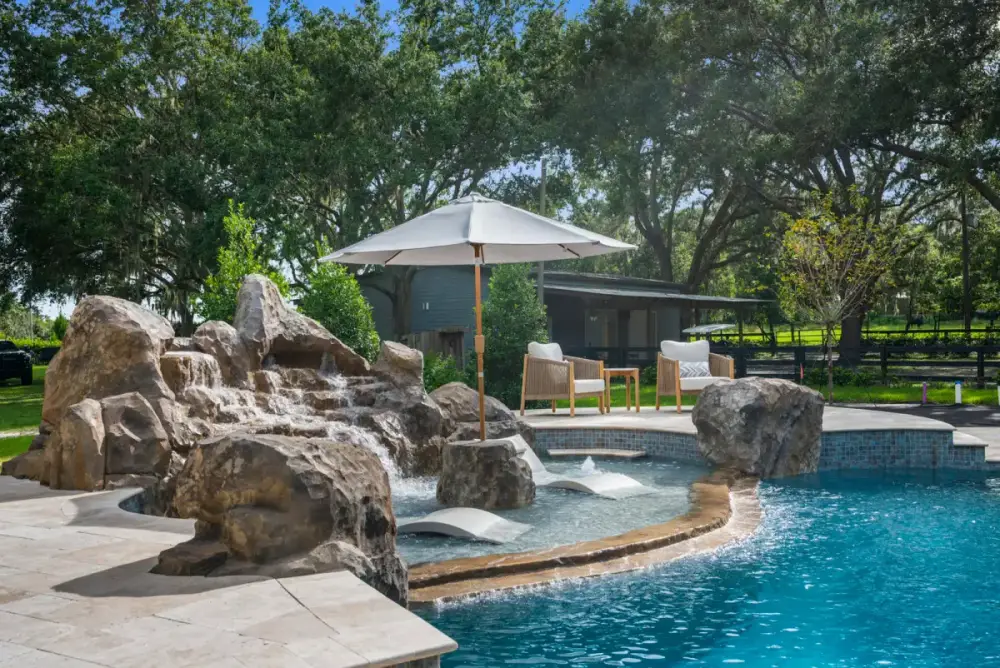 Naturalistic custom pool featuring a built-in rock waterfall, large shaded umbrella, and surrounding tropical landscaping.