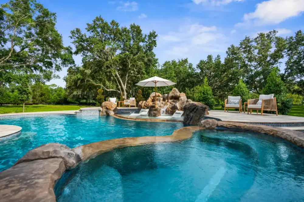 Spacious custom pool featuring a shaded Baja shelf with umbrella, surrounded by natural stone and lush trees for a resort-style feel.