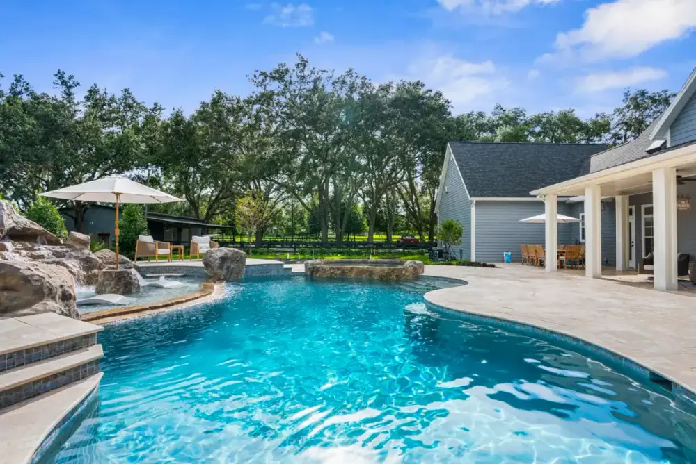 Crystal-clear custom pool featuring a sun shelf entry, bordered by a modern patio and surrounded by lush greenery.