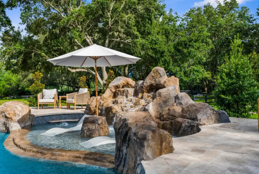 Inviting pool entryway with a sun shelf and custom rock waterfall, enhanced by integrated landscaping and a shaded lounging area.