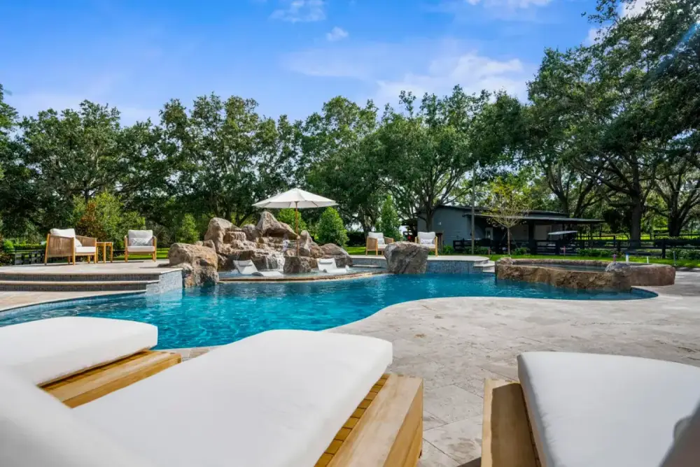 Low-angle view from in-pool loungers facing a custom pool with clear blue water, surrounded by trees and outdoor seating.