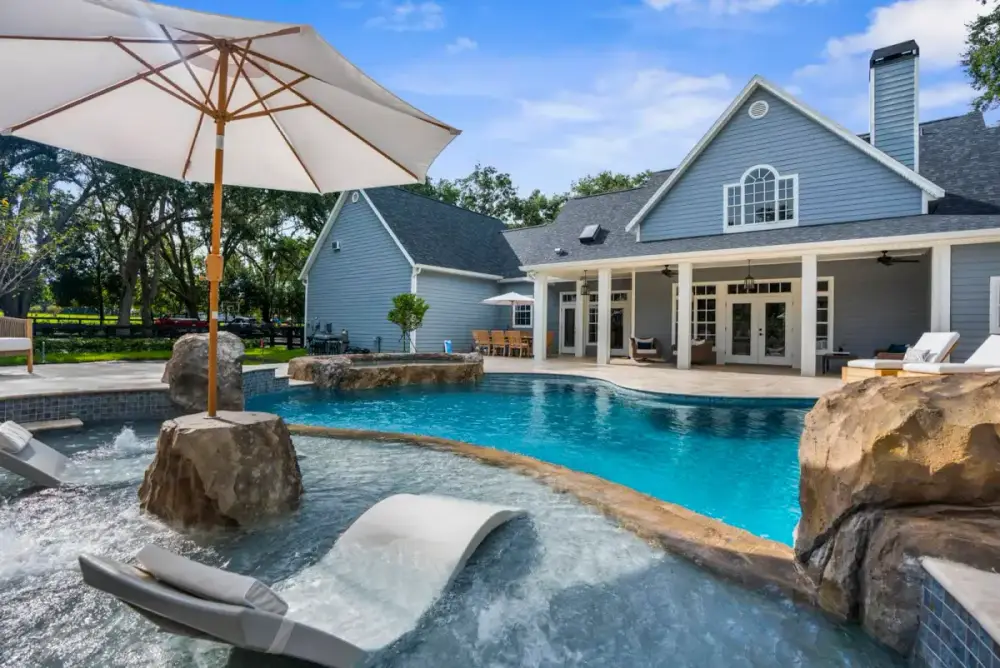 Shaded in-pool loungers set on a baja shelf beneath an umbrella, overlooking a luxury custom pool with rock accents and home exterior in view.