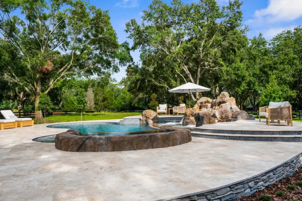 Spacious patio area around a custom pool with a built-in elevated spa and curved deck design, nestled in a wooded yard.